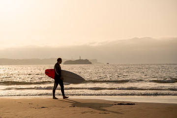 Naklejka premium Surfer walking with surfboard on the beach at sunset or sunrise. Vacation and adventure lifestyle