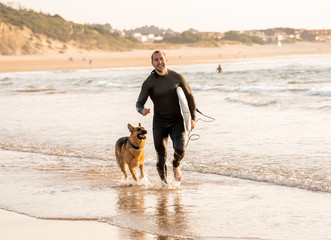 Surfer man with his dog german shepherd and surfboard playing and surfing on the beach