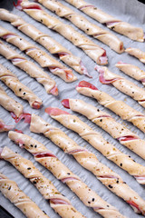 Sticks of raw puff dough with bacon sprinkled with sesame seeds on the oven tray ready to be baked