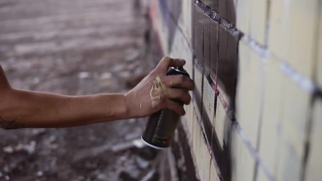 Close-up of artists hand dirty in paint applying spray drawing a black colour on a street building wall. Action. Stained fingers of artist hold spray can with colored paint on concrete wall. Street