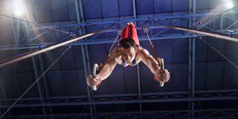Male athlete doing a complicated exciting trick on gymnastics rings in a professional gym. Man perform stunt in bright sports clothes - obrazy, fototapety, plakaty