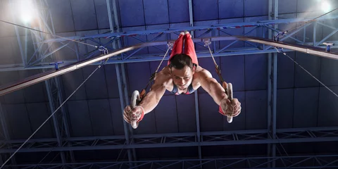 Poster Im Rahmen Male athlete doing a complicated exciting trick on gymnastics rings in a professional gym. Man perform stunt in bright sports clothes © Alex