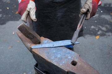 At the smithy workshop. Blacksmith’s hands holding forceps and a hammer forging a metal billet, blade of a knife, on an anvil