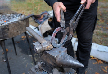 At the smithy workshop. Blacksmith’s hands holding forceps and a hammer forging a metal billet, horseshoe, on an anvil
