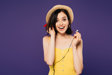 excited smiling pretty girl in straw hat putting on earphones isolated on purple