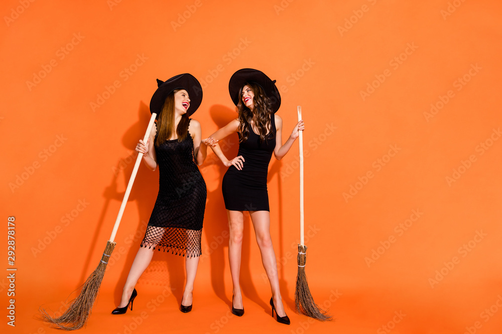 Poster Photo of two cruel witch ladies holding brooms evil laughing wear black dresses and wizard hats isolated orange color background