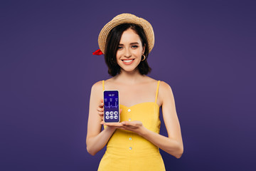 smiling pretty girl in straw hat holding smartphone with healthcare app isolated on purple