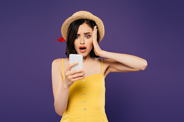 shocked pretty girl in straw hat using smartphone isolated on purple