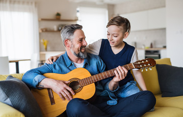 Mature father with small son sitting on sofa indoors, playing guitar.