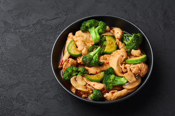 Hunan Chicken in black bowl at dark slate background. Hunan Chicken is chinese or indo-chinese cuisine takeaway dish with broccoli, zucchini, shiitake mushrooms and hunan sauce