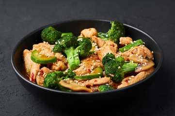 Hunan Chicken in black bowl at dark slate background. Hunan Chicken is chinese or indo-chinese cuisine takeaway dish with broccoli, zucchini, shiitake mushrooms and hunan sauce