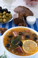 Traditional Russian soup Solyanka cooked with meat, sausages, salted cucumbers on wite bowl. Black bread, lemon, olived and tomatoes on color blue background, homemade food.