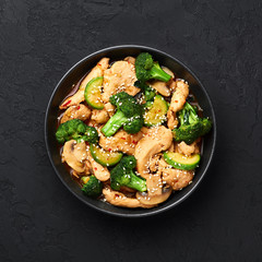 Hunan Chicken in black bowl at dark slate background. Hunan Chicken is chinese or indo-chinese cuisine takeaway dish with broccoli, zucchini, shiitake mushrooms and hunan sauce. Top view