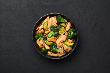 Hunan Chicken in black bowl at dark slate background. Hunan Chicken is chinese or indo-chinese cuisine takeaway dish with broccoli, zucchini, shiitake mushrooms and hunan sauce. Copy space. Top view