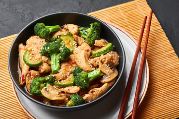 Hunan Chicken in black bowl at dark slate background. Hunan Chicken is chinese or indo-chinese cuisine takeaway dish with broccoli, zucchini, shiitake mushrooms and hunan sauce