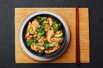 Hunan Chicken in black bowl at dark slate background. Hunan Chicken is chinese or indo-chinese cuisine takeaway dish with broccoli, zucchini, shiitake mushrooms and hunan sauce. top view