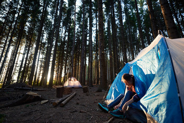 Morning camping in the forest. Young woman sitting in blue tourist tent, tying the shoe laces, getting ready to hike. On background forest, woods and white tent. Tourism active lifestyle concept