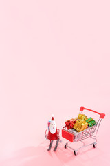 Christmas shopping concept, mini red shop cart trolley with Santa Claus toy and gift box isolated on pale pink background, blank copy space, close up