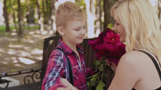 Beautiful little boy gives a bouquet of flowers to his beloved mother in the park on a bench.