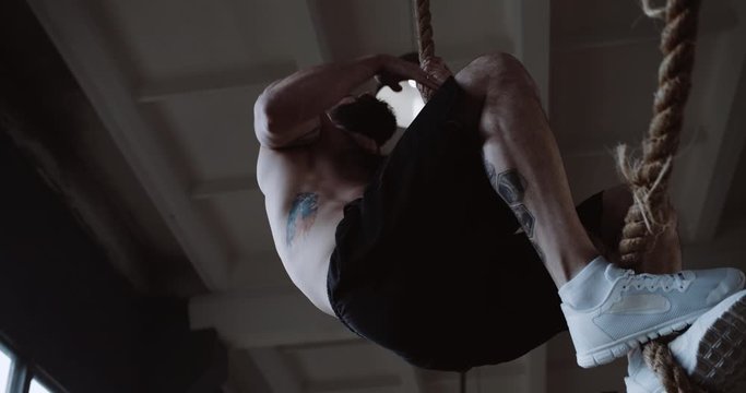 Young muscular athletic Caucasian man climbing rope, doing hardcore functional workout training in large gym slow motion