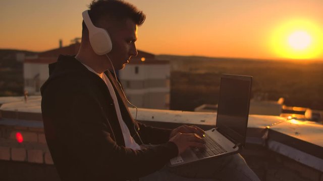 A man freelancer in headphones standing on the roof at sunset writes on the keyboard code pages. Little business. Listen to music and work at the computer enjoying the beautiful view from the roof