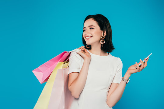 smiling elegant woman in dress with shopping bags and smartphone looking away isolated on blue