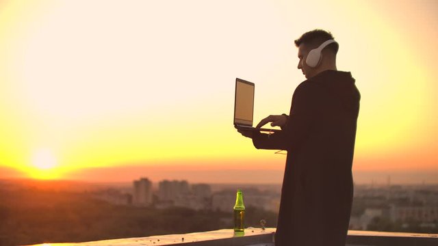 A man freelancer in headphones standing on the roof at sunset writes on the keyboard code pages. Little business. Listen to music and work at the computer enjoying the beautiful view from the roof