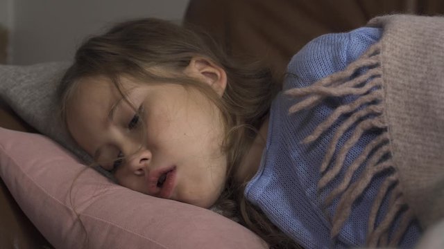 Sick young caucasian girl coughing while lying under blanket at home. The child has fever. Concept of health, illness, sickness, common cold, treatment