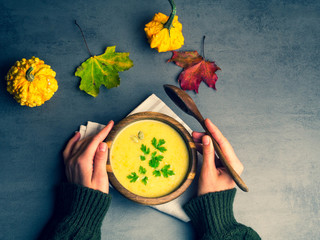 Bowl of vegetarian autumn pumpkin carrot soup, served with parsley, seeds, on textile on dark gray kitchen table with colorful foliage. Female hands. Top view with space