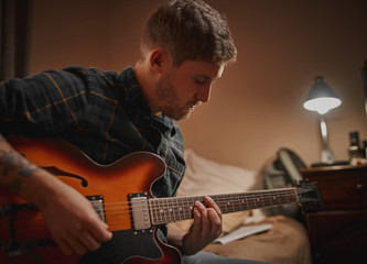 Close-up of a young man practicing playing guitar at evening in his home