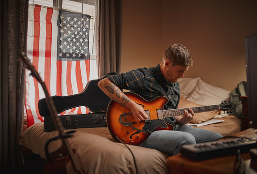 Young Guitarist Man At Home Sitting On Bed In Front Of Window With Usa Flag Playing Guitar