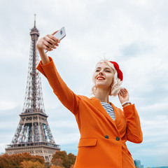 Happy smiling tourist girl makes selfie in Paris with view of Eiffel tower. Lady wearing red beret,...