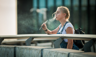 Young woman outdoor vaping e-cigarette on modern city buildings background