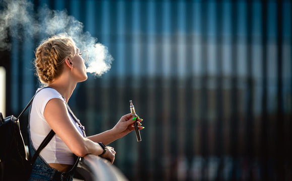 Young woman outdoor vaping e-cigarette on modern city buildings background