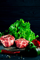 Two juicy steaks lie on a wild stone next to red and green chili peppers and a fresh green salad on a black wooden table