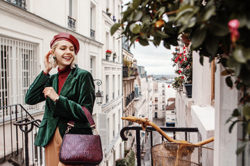 Outdoor autumn portrait of young happy smiling fashionable lady wearing trendy leather beret, turtleneck, green velour blazer, earrings, holding textured bag, posing in street of Paris. Copy space