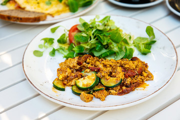 Vegan breakfast prepared with scrambled tofu and eggs, cooked with dried tomatoes and zucchini served with fresh lettuce and cucumber in white ceramic vintage plate