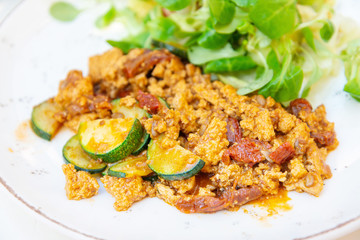 Vegan breakfast prepared with scrambled tofu and eggs, cooked with dried tomatoes and zucchini served with fresh lettuce and cucumber in white ceramic vintage plate
