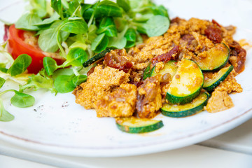 Vegan breakfast prepared with scrambled tofu and eggs, cooked with dried tomatoes and zucchini served with fresh lettuce and cucumber in white ceramic vintage plate