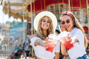 Two young beautiful girls have fun in an amusement park. Drink lemonade and laugh