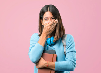 Young student girl with blue sweater and headphones smiling a lot while covering mouth on pink background