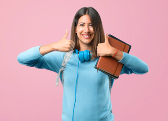 Young student girl with blue sweater and headphones giving a thumbs up gesture and smiling because has had success on pink background
