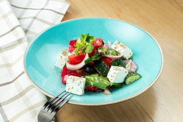 Greek salad with olives, tomatoes, feta cheese, onions in blue bowl on wooden background. Close up. Selective focus