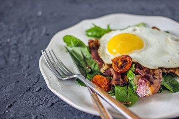 Appetizing breakfast with fried eggs and salad leaves. From above delicious meal with bacon egg tomatoes and green leaves served on white plate over gray textured background