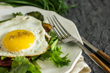 Appetizing breakfast with fried eggs and salad leaves. From above delicious meal with bacon egg tomatoes and green leaves served on white plate over gray textured background