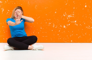 young blonde woman covering face with hand and putting other hand up front to stop camera, refusing...