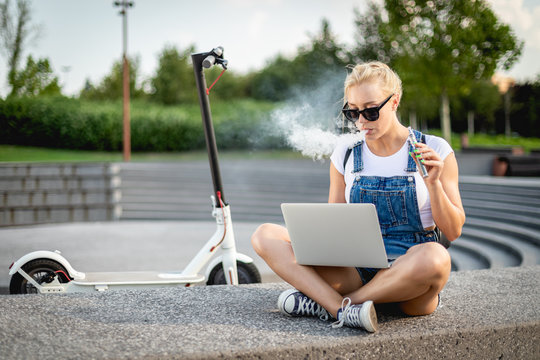 Modern woman smoking e-cigarette using her laptop computer while sitting at street with electric scooter
