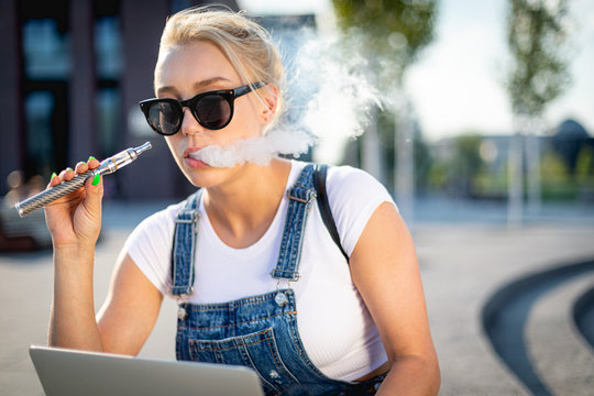 Modern woman smoking e-cigarette and using her laptop computer while sitting at street