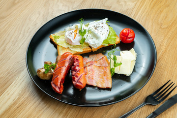 Close up on fried eggs with bacon, sausages, dor blue cheese on a stylish black plate. Food for breakfast. Restaurant Serving. Wooden background