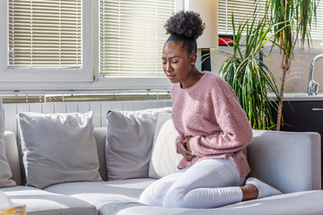 African American Woman in painful expression holding hands against belly suffering menstrual period pain, lying sad on home bed, having tummy cramp in female health concept - obrazy, fototapety, plakaty
